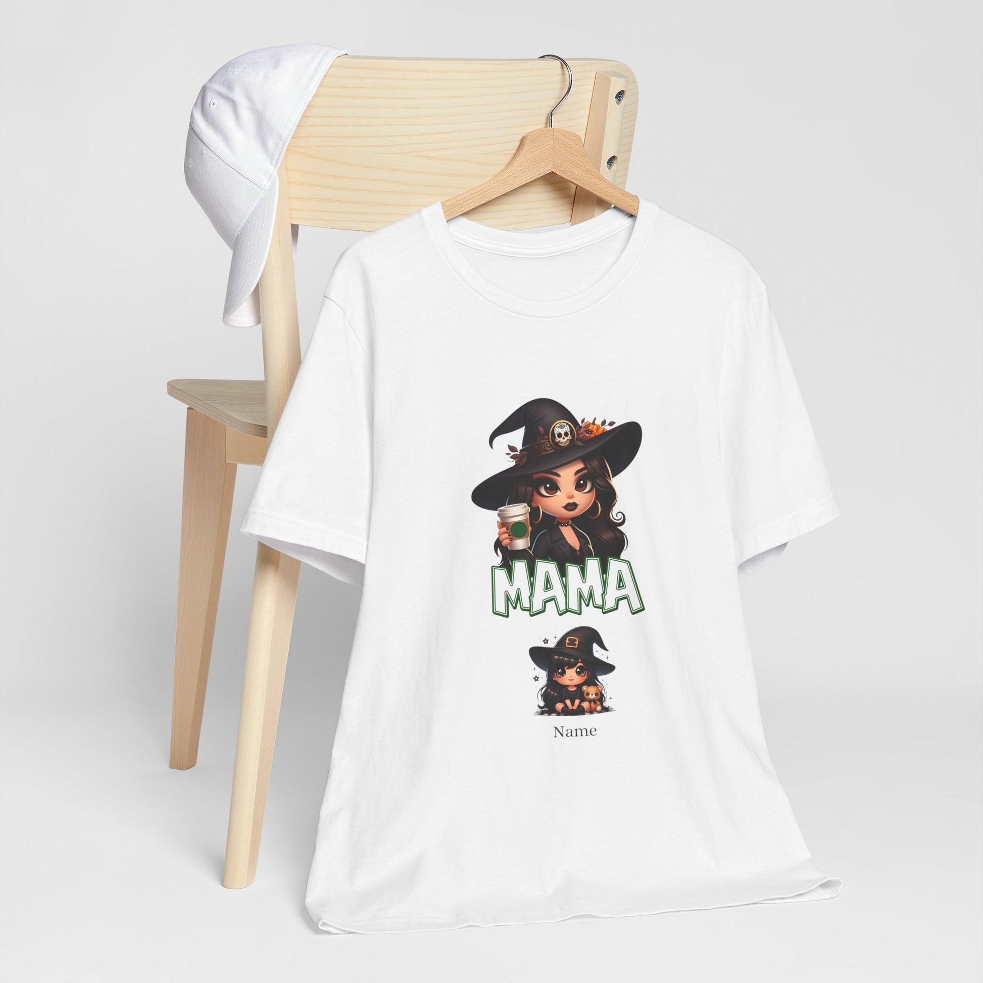 Displayed on a wooden chair is the Unisex Jersey Cute Witch Personalized Mama Short Sleeve Tee by Printify. This white T-shirt features a customizable witch-themed graphic design with a cartoon witch holding a cup, and the word "MAMA" in bold green letters below. A white cap rests on the chair's backrest.