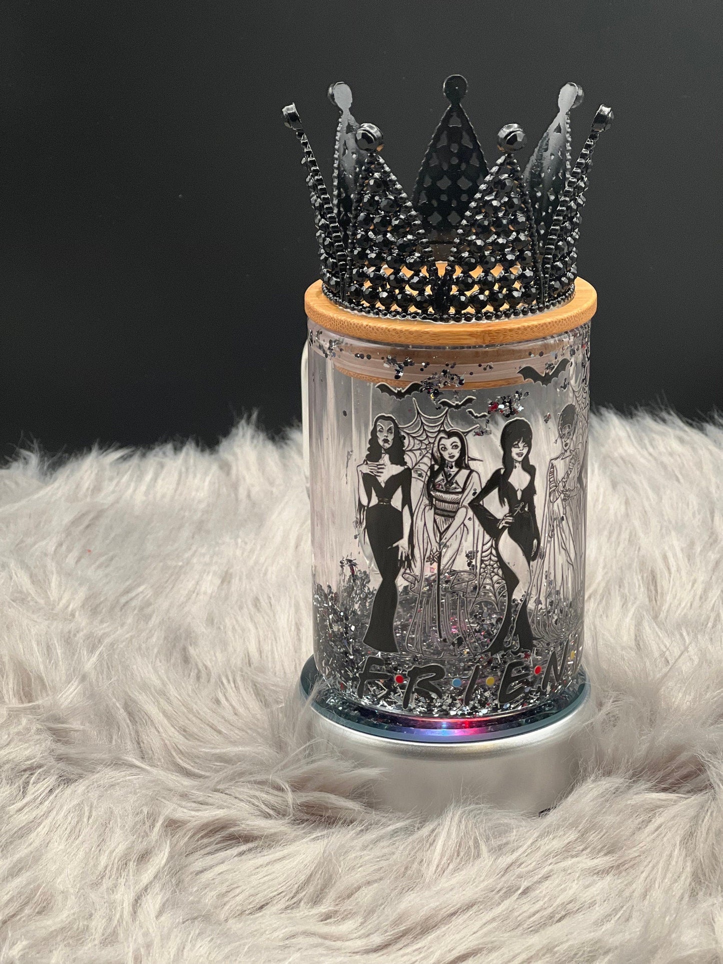 A 15oz Glass Mug by Just For Lookz, featuring detailed images of Elvira, Bride of Frankenstein, and Morticia, rests on a fluffy white surface. The mug is topped with a bamboo lid that adds to its Gothic Elegance. Soft, diffused lighting highlights the intricate design and spooky decor.