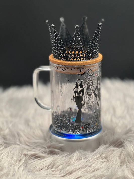 A 15oz Glass Mug with Elvira, Bride of Frankenstein, and Morticia Design by Just For Lookz is placed on a fluffy gray surface. The mug, featuring elegant illustrations of these iconic gothic women, is topped with a black bejeweled crown and accompanied by a snow globe and bamboo lid, adding an artistic and gothic decor touch to the regal setup.