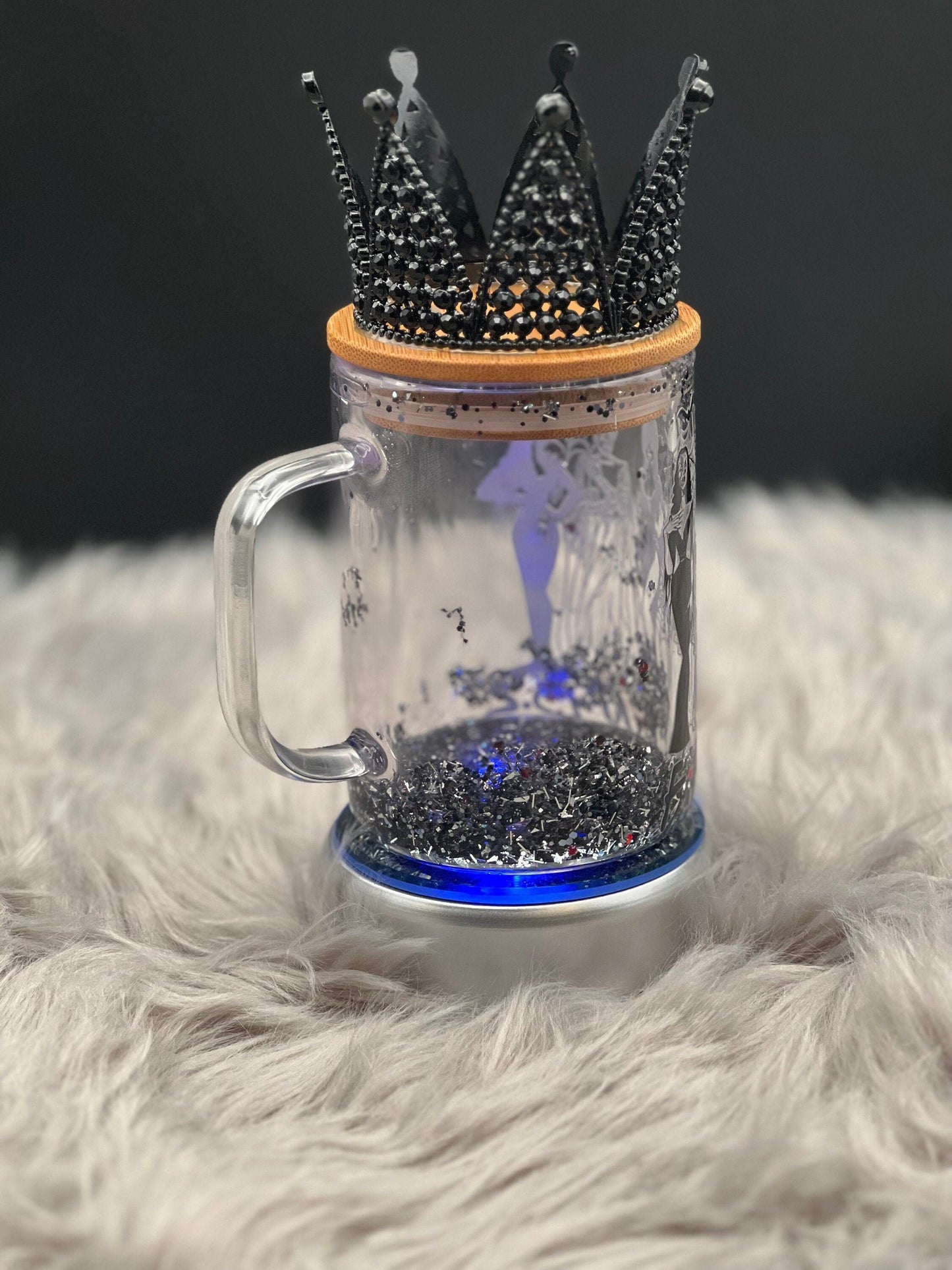 A 15oz glass mug from Just For Lookz, featuring intricate silhouette designs of Elvira, Bride of Frankenstein, and Morticia, rests atop a fluffy gray surface. This exquisite piece of gothic decor is filled with sparkling black glitter and adorned with an ornate black crown on its bamboo lid. Eerie blue light glows from the base, adding a touch of horror-themed elegance to this stunning snow globe mug.