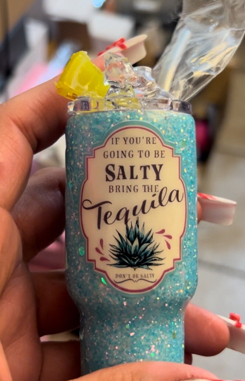 A hand holds a blue glittery Delightful 2oz Mini Glittered Tumbler from Just For Lookz, labeled "If you're going to be salty, bring the tequila" with an image of a tequila plant. The tumbler has an open top with faux ice cubes and lemon coming out. The person's nails are decorated with pink and white designs.