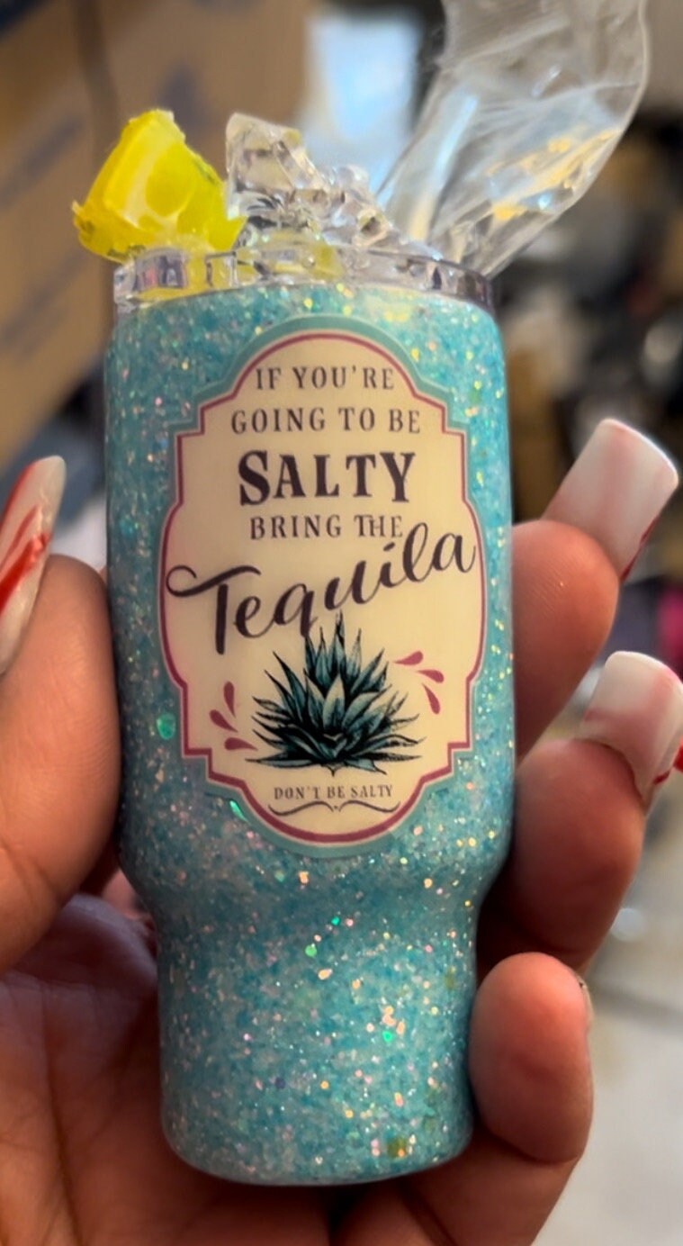 A person with long, manicured nails holds Just For Lookz's Delightful 2oz Mini Glittered Tumbler, adorned with the humorous quote "If you're going to be salty, bring the tequila" above an illustration of an agave plant. The stainless steel tumbler contains faux ice cubes and a faux lemon slice inside the opening.