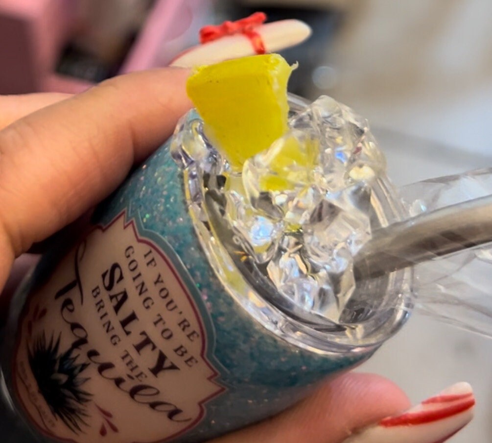 Close-up of a person holding a blue bottle with crushed ice, a lemon wedge, and a straw. The bottle bears the text "If you're going to be salty, bring the tequila." Displaying their red-tipped nails and a red string tied around their finger, they show off their Just For Lookz Delightful 2oz Mini Glittered Tumbler featuring faux ice cubes and a custom lid with lemon.