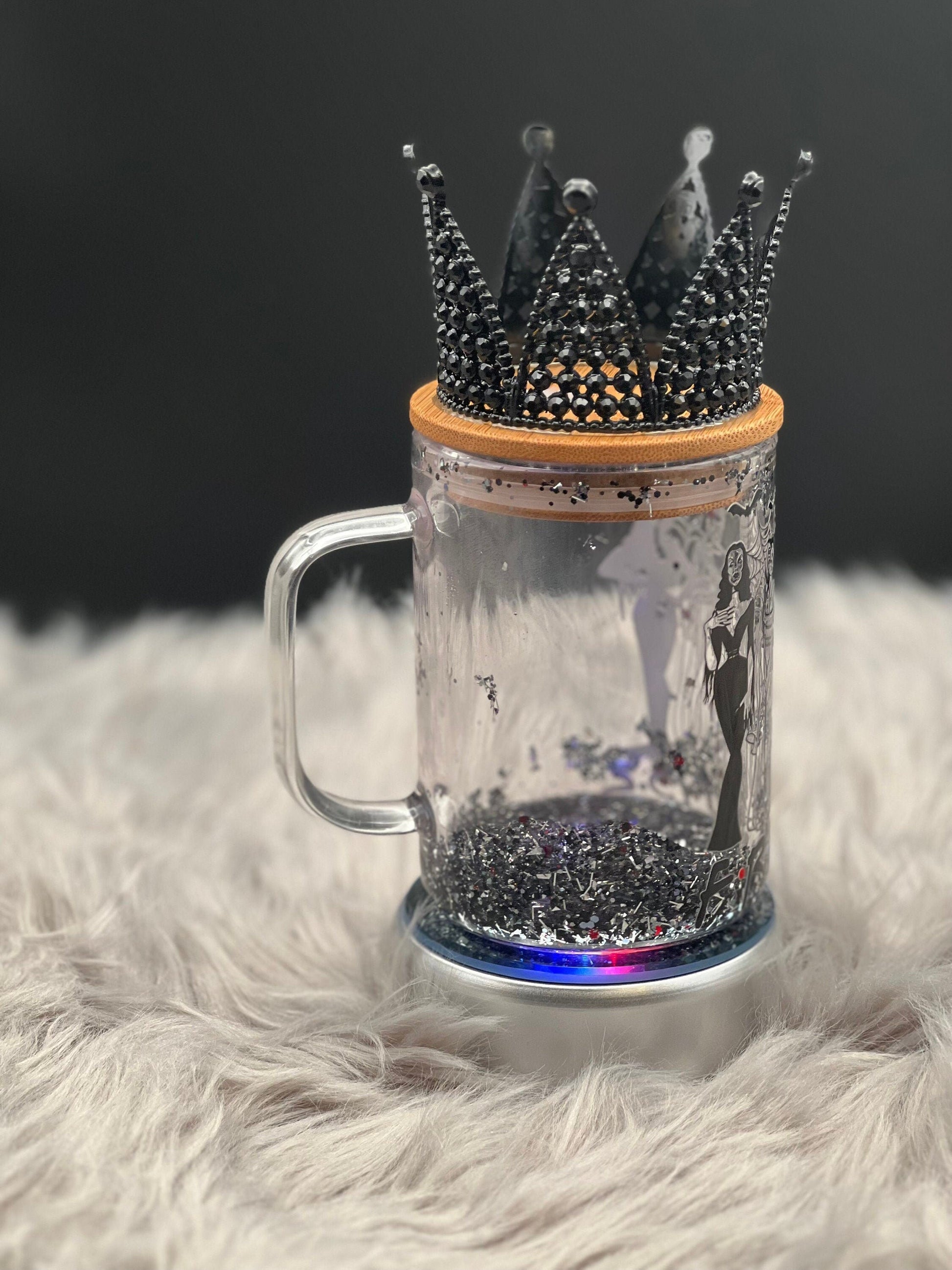 A 15oz glass mug from Just For Lookz, featuring an Elvira, Bride of Frankenstein, and Morticia design, is topped with a bamboo lid adorned with a black jeweled crown. This Gothic Elegance mug sits on a fluffy white surface against a dark, blurred background. The intricate and sparkling details of the crown make it stand out beautifully.