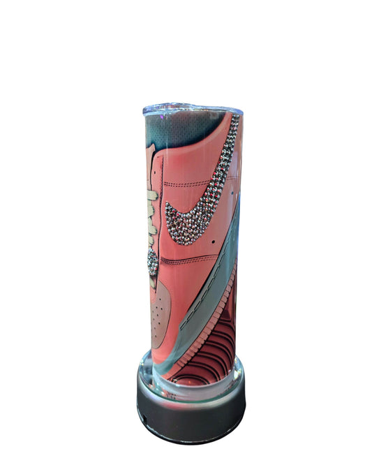 A cylindrical display case showcasing the "20oz Stainless Steel Rhinestone Tumbler Nike Inspired" from Just For Lookz. The tumbler is adorned with rhinestone decorations on the Nike swoosh and positioned upright to provide a clear view of its side profile. The base of the display case is metallic.