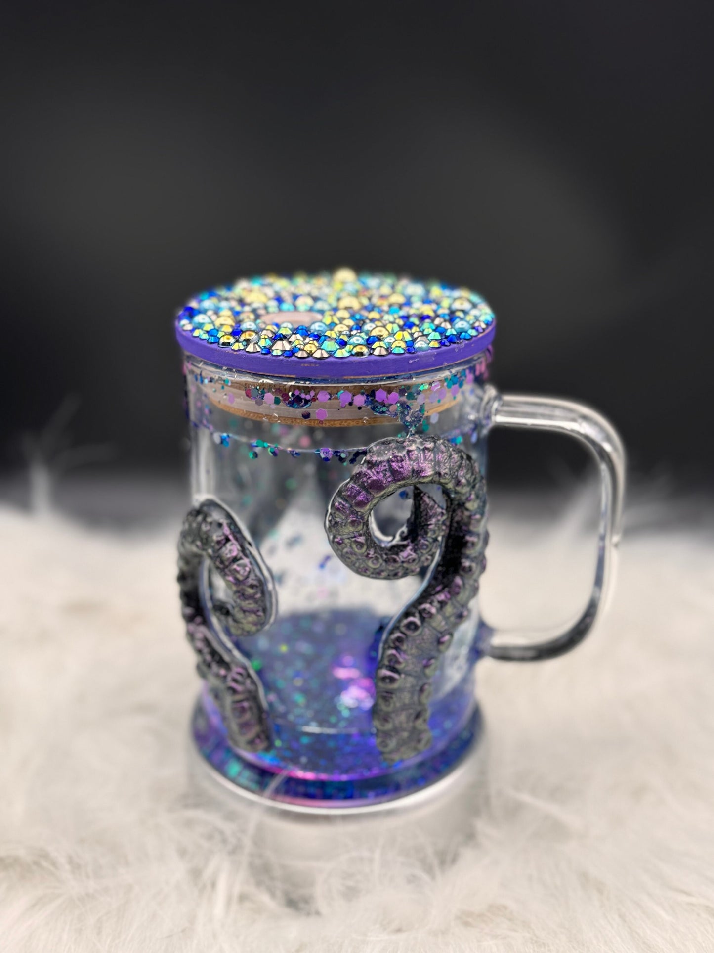 The **Discover the Enchantment: 3D Octopus Tentacle Ursula Glass Mug** by Just For Lookz is a clear glass mug with a handle, beautifully adorned with purple sparkly octopus tentacles wrapped around its sides. The lid is decorated with colorful beads and sequins, making it perfect for your undersea adventures or as an Ocean Enthusiast Gift. This alluring Ursula Glass Mug rests elegantly on a white, fluffy surface with a dark, blurred background.