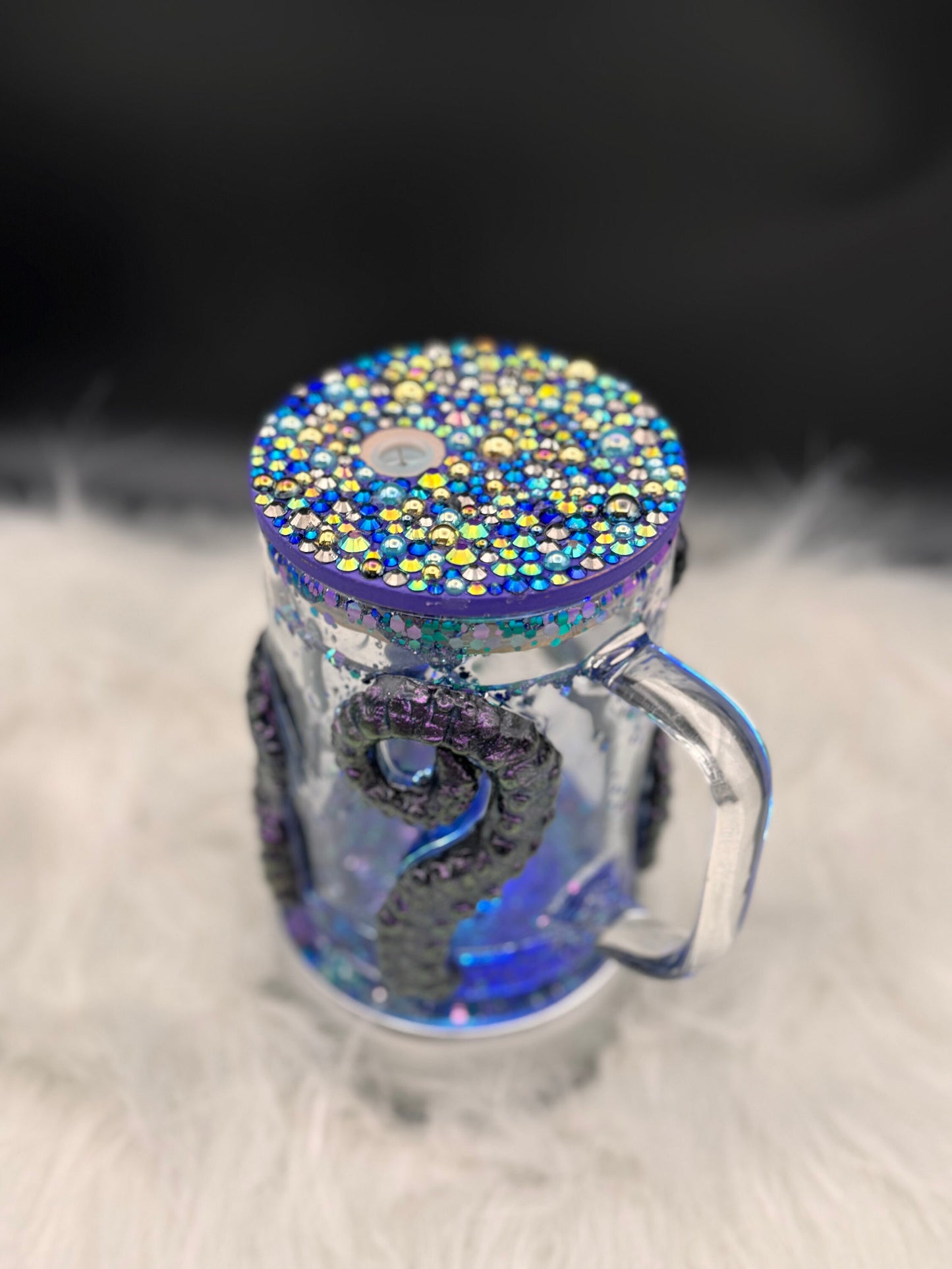 A clear Discover the Enchantment: 3D Octopus Tentacle Ursula Glass Mug from Just For Lookz, featuring a handle and a colorful, gem-studded lid, sits on a fuzzy white surface. Lining the inside of the tumbler are blue tentacles with suction cups, making it perfect for any ocean enthusiast.