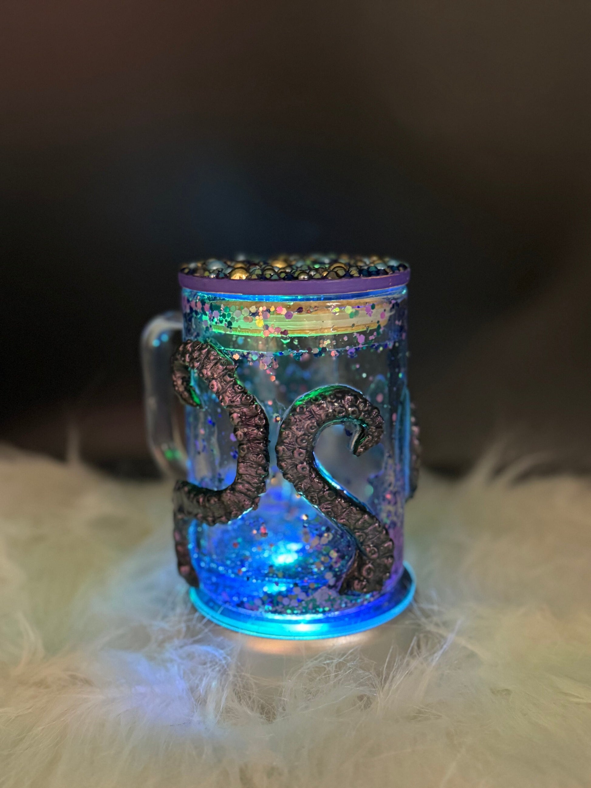 The "Discover the Enchantment: 3D Octopus Tentacle Ursula Glass Mug - Perfect for Your Undersea Adventures!" by Just For Lookz, featuring transparent glass with dark, tentacle-like designs wrapping around it, rests on a soft, white, furry surface. The mug contains colorful speckles and is illuminated with glowing blue light, casting an ethereal effect against a dark background.