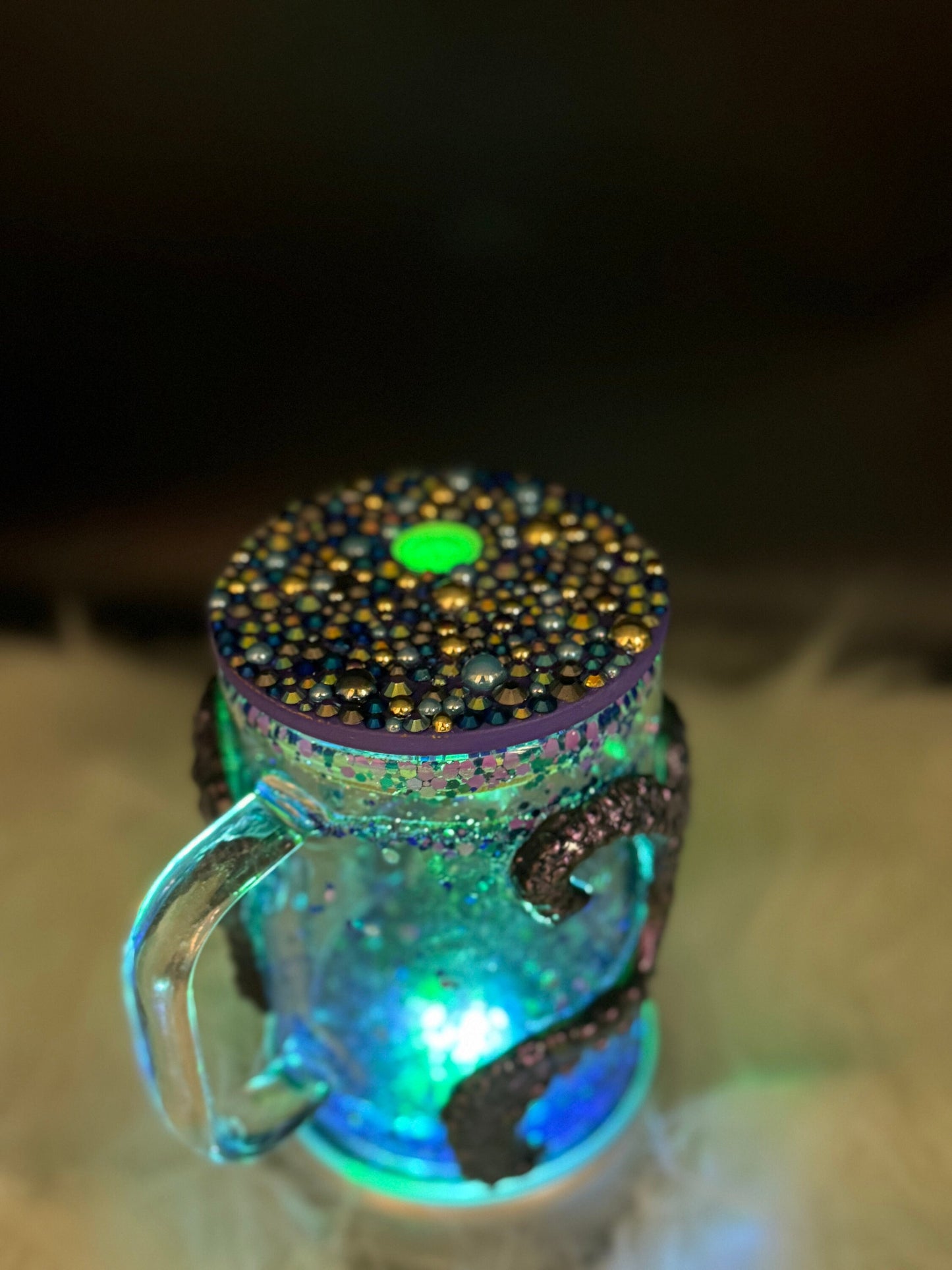 A glass mug, perfect for the ocean enthusiast, is adorned with intricate decorations made of beads, sequins, and shells. Featuring a glowing light inside and a lid covered in dense multicolored beads with a prominent green one near the center, this "Discover the Enchantment: 3D Octopus Tentacle Ursula Glass Mug" by Just For Lookz is truly captivating.