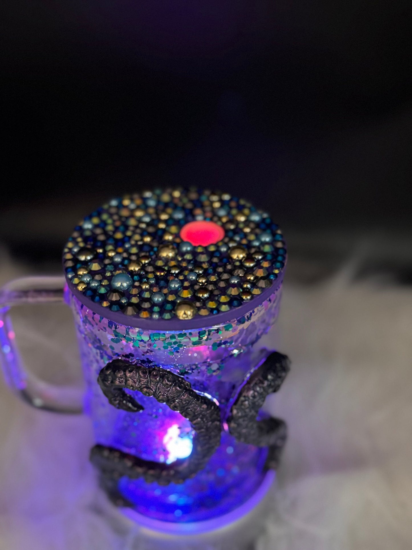 The Discover the Enchantment: 3D Octopus Tentacle Ursula Glass Mug from Just For Lookz features a decorative glass body illuminated from within. It is adorned with intricate glittery designs and bold, dark embellishments of the number "24" against a blurred, dark background. The textured blue and gold lid has a striking pinkish centerpiece that completes its enchanting look.